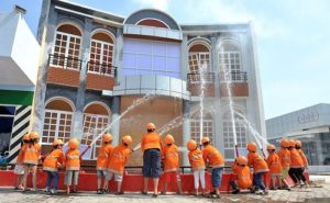 Nhóm trẻ mặc áo cam và mũ bảo hộ đứng quanh đài phun nước trước mặt tiền mô phỏng tại KizCity Quận 4