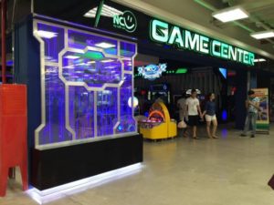Lối vào Game Center trong khu vui chơi Apple Kids Clup, bảng hiệu neon xanh, dãy máy arcade và người ra vào