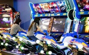 Hai cô gái chơi máy đua xe mô tô trong khu vui chơi arcade, ghế xanh và màn hình game sáng
