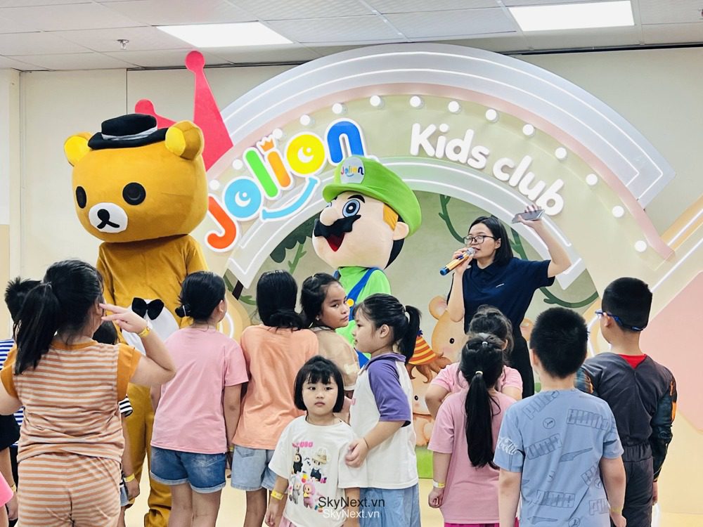 Trẻ em tụ tập trước hai mascot gấu và nhân vật đội mũ xanh cùng nữ hướng dẫn
