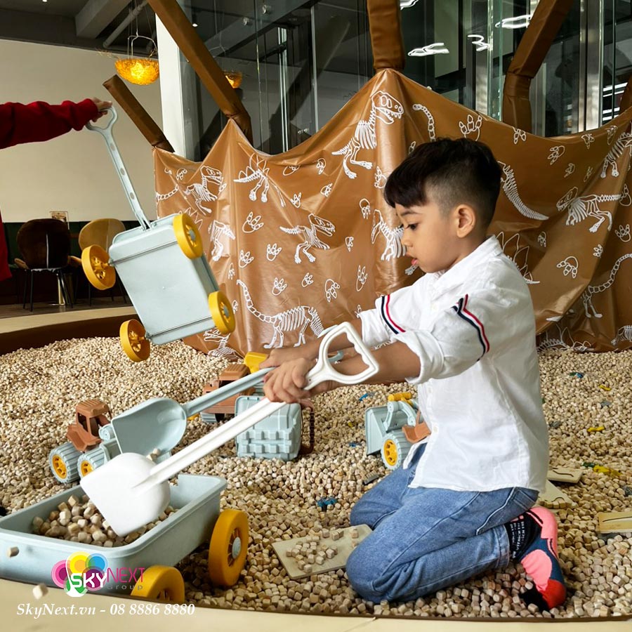 Bé trai quỳ xúc hạt nhựa bằng xẻng đồ chơi cạnh xe kéo